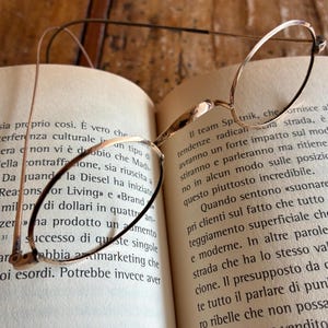 May include: Close-up of gold-rimmed eyeglasses resting on an open book. The book's pages are filled with Italian text. The glasses have a thin frame and oval lenses, with a classic, vintage aesthetic.
