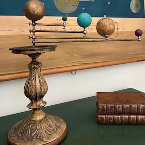 May include: A mechanical orrery model featuring a textured gold sphere, a small dark sphere, a teal sphere, a larger patterned globe, a tiny white sphere, and a small purple sphere, all mounted on a decorative, ornate gold-toned metal pedestal base.