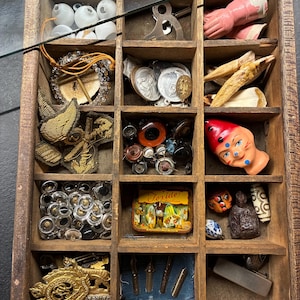 Puede incluir: Una bandeja de madera dividida en compartimentos con varios objetos vintage. Contiene cuentas de vidrio, objetos metálicos, una pequeña cabeza de muñeca, botones y una lata con la palabra "Jeride". Los objetos están dispuestos en compartimentos individuales.