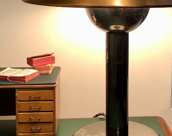 May include: A vintage brass and black desk lamp with a round shade and a round base. The lamp is on a green desk with a stack of books.