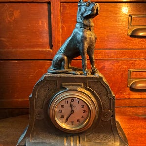 Puede incluir: Un reloj antiguo con una estatua de perro de color bronce sobre una base decorativa. La esfera del reloj tiene números y manecillas negras, con un diseño circular. El perro está sentado y mirando hacia arriba.
