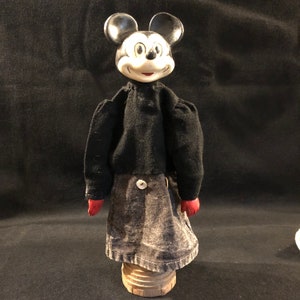Puede incluir: Un t&iacute;tere de mano vintage de Mickey Mouse con un cuerpo de fieltro negro, guantes de fieltro rojo y una cabeza de porcelana negra y blanca. El t&iacute;tere est&aacute; de pie sobre una base de madera.