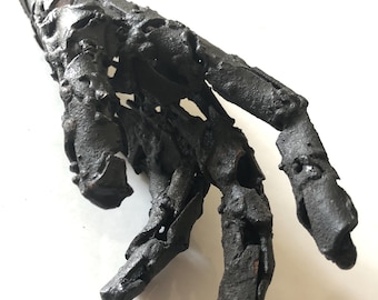 Könnte beinhalten: Eine dunkle, texturierte Skulptur einer menschlichen Hand und eines Unterarms aus geschweißtem Metall. Die Oberfläche weist ein raues, unebenes Finish mit sichtbaren Schweißnähten und Lücken zwischen den Segmenten auf.