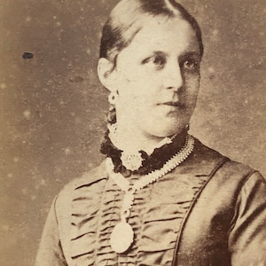 May include: A sepia toned portrait of a woman wearing a dark dress with a ruffled neckline and a pearl necklace with a locket.