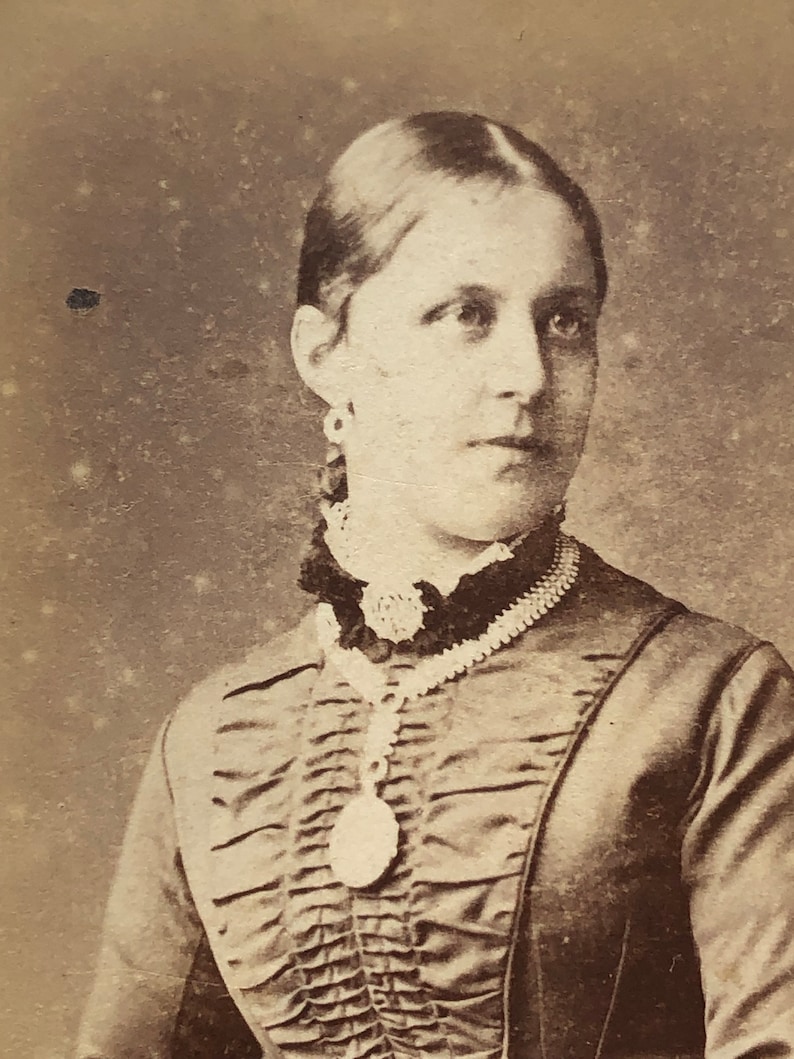 May include: A sepia toned portrait of a woman wearing a dark dress with a ruffled neckline and a pearl necklace with a locket.