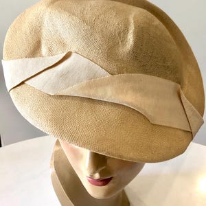 May include: A beige straw hat with a wide brim and a white ribbon. The hat is angled slightly to the side.