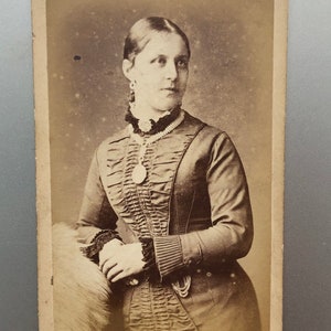 May include: A black and white portrait photograph of a woman wearing a dark dress with a ruffled neckline and a pearl necklace. The photo is framed with a white border and has the text 'REES, & C., 298, CLAPHAM ROAD S.W.' at the bottom.