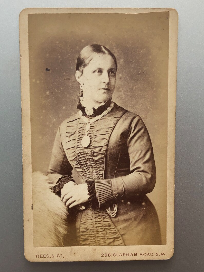 May include: A black and white portrait photograph of a woman wearing a dark dress with a ruffled neckline and a pearl necklace. The photo is framed with a white border and has the text 'REES, & C., 298, CLAPHAM ROAD S.W.' at the bottom.