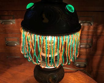 May include: A vintage lamp with a beaded shade and green glass accents. The lamp base is decorated with a floral design and a portrait.