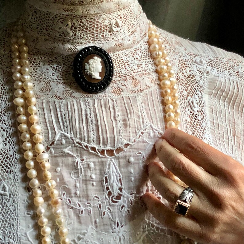 May include: A white lace blouse with a cameo brooch and a pearl necklace. The blouse has a high neckline and a gathered bodice.
