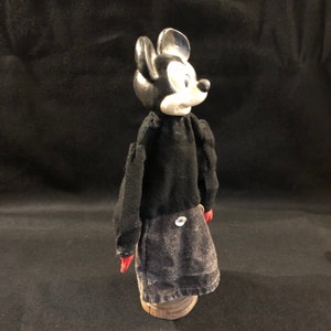 Puede incluir: Un t&iacute;tere vintage de Mickey Mouse en blanco y negro con guantes rojos y una camisa negra. El t&iacute;tere lleva una falda vaquera con un bot&oacute;n en la espalda. El t&iacute;tere est&aacute; de pie sobre una base de madera.