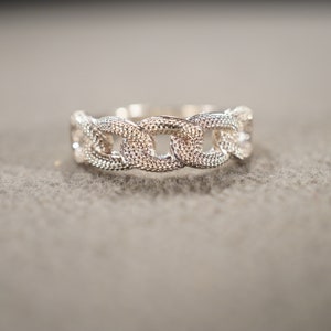 May include: A silver ring with a chain-link design. The ring is made of multiple interconnected oval-shaped links.