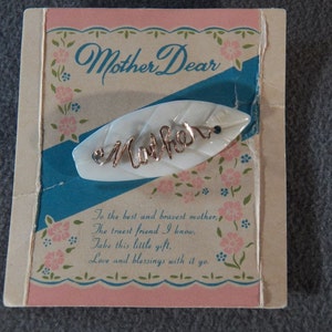May include: A white mother's day pin with a gold inscription that reads "Mother" on a blue and white background. The pin is on a card with the text "Mother Dear" and a poem that reads "To the best and bravest mother, The truest friend I know, Take this little gift, Love and blessings with it yo."