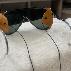 May include: Black sunglasses with brown leather side shields and a silver metal frame. The sunglasses have a black elastic strap.