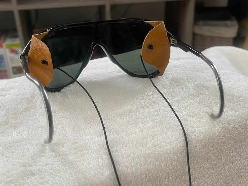 May include: Black sunglasses with brown leather side shields and a silver metal frame. The sunglasses have a black elastic strap.