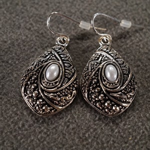 May include: A pair of silver earrings with a teardrop shape and a white pearl in the center. The earrings have a detailed, ornate design.