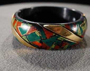 May include: A black bangle bracelet with a geometric pattern in shades of green, red, and gold. The bracelet is made of plastic and has a shiny finish.