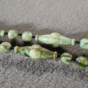 May include: A green and gold glass bead necklace with a black cord. The beads are round and have a marbled pattern.