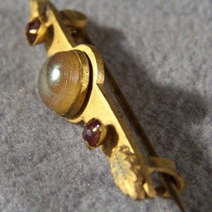 May include: A gold-toned brooch with a large iridescent stone and two smaller red stones. The brooch has a decorative design and a pin on the back.