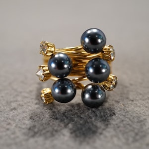 May include: A gold-toned ring featuring a spiral design with six dark gray pearls and six clear, sparkling accent stones. The ring's design is modern and eye-catching, suitable for everyday wear or special occasions.