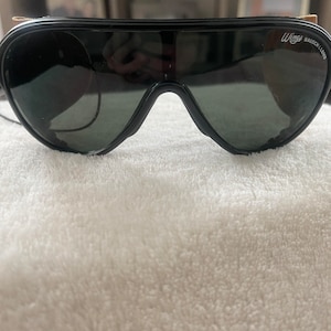 May include: Black aviator sunglasses with a brown leather strap and the text "Wings BAUSCH & LOMB" on the right lens.