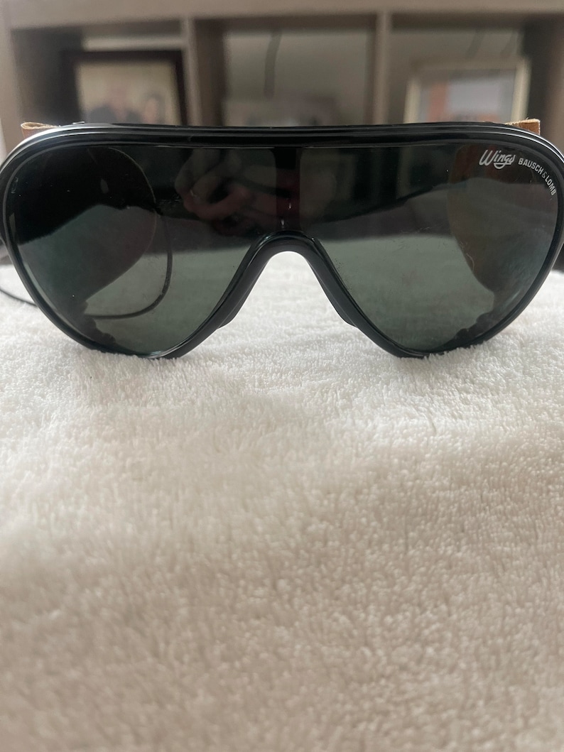 May include: Black aviator sunglasses with a brown leather strap and the text "Wings BAUSCH & LOMB" on the right lens.