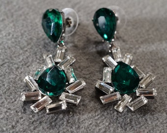 May include: A pair of dangle earrings with a teardrop-shaped green gemstone surrounded by smaller clear rhinestones.