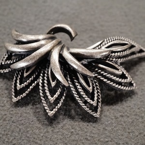 May include: A silver-toned metal hair clip with a floral design. The clip has a swirl design in the center and multiple layers of delicate, intricate details.