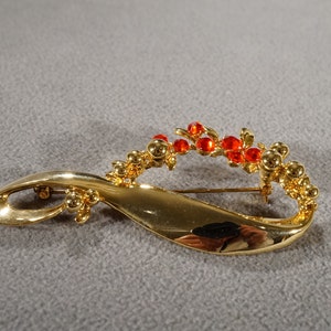 May include: Gold-toned brooch with a stylized, abstract design featuring a curved, flowing shape and a cluster of red rhinestones.