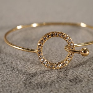 May include: A gold bangle bracelet with a circle charm set with clear rhinestones.