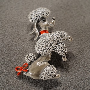May include: A silver poodle brooch with white and gray detailing. The poodle has a red bow around its neck.