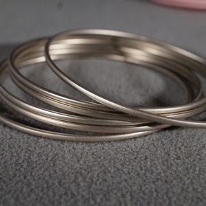 May include: A set of five silver-toned metal bangles stacked on top of each other. The bangles are smooth and shiny.