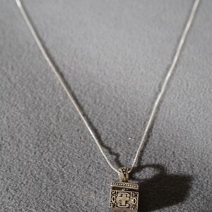 May include: A silver chain necklace with a small silver pendant shaped like a box with a cross on the front.