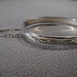 May include: A silver bangle bracelet with a decorative etched design. The bracelet has a chain attached to it.