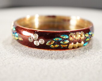 May include: A red bangle bracelet with gold and iridescent blue and green enamel accents. The bracelet is decorated with clear rhinestones.