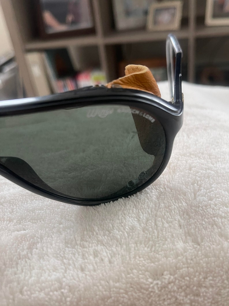 May include: Black sunglasses with a brown leather nose pad and the text "Wayfarer" on the frame.