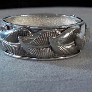May include: Silver tone bangle bracelet with a leaf design. The bracelet is made of metal and has a hinged clasp.