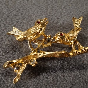 May include: Gold-tone brooch featuring two birds perched on a branch with clear rhinestone accents. The birds have red eyes.