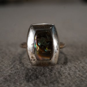 May include: A silver ring with a rectangular abalone shell inlay. The shell has iridescent colors of green, blue, and gold.