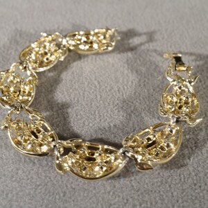 May include: A gold-tone bracelet with a delicate, intricate design. The bracelet is made up of a series of connected oval-shaped links, each with a detailed floral pattern. The bracelet has a clasp closure.