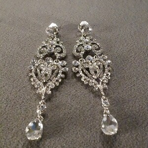 May include: A pair of silver dangle earrings with clear crystal embellishments. The earrings feature a delicate, intricate design with multiple layers of crystals.