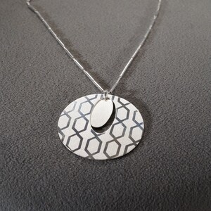 May include: A silver necklace with a geometric pendant. The pendant is a circle with a smaller oval cutout in the center. The circle has a pattern of hexagons.