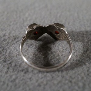 May include: A silver ring with two red gemstones set in the center of the band. The ring is designed in a heart shape.