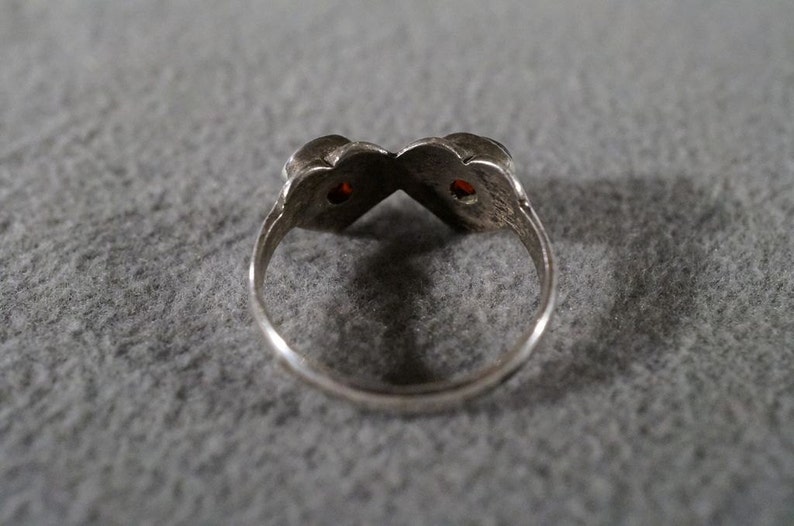May include: A silver ring with two red gemstones set in the center of the band. The ring is designed in a heart shape.