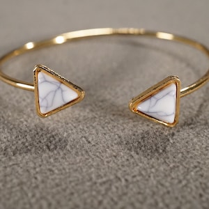 May include: Gold-tone open cuff bracelet with white and gray triangular stone accents. The bracelet features a minimalist design with a polished finish. The triangular stones are set in gold-tone frames.