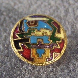 May include: A round, gold-rimmed button with a colorful geometric design. The design features a blue, yellow, green, red, and black pattern.