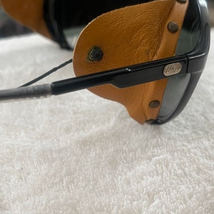 May include: Black sunglasses with brown leather side shields and a black strap. The sunglasses have a silver logo that says "Wiley X" on the right side.