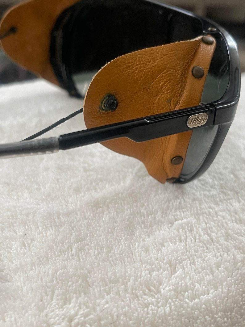 May include: Black sunglasses with brown leather side shields and a black strap. The sunglasses have a silver logo that says "Wiley X" on the right side.