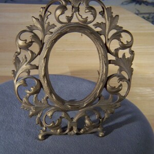 May include: A gold-toned metal picture frame with an ornate design featuring a floral motif. The frame has an oval opening and stands on a base.