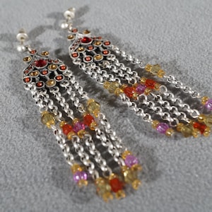 May include: A pair of silver dangle earrings with a multi-colored beaded chain. The earrings feature a silver and red jeweled top with a decorative design.